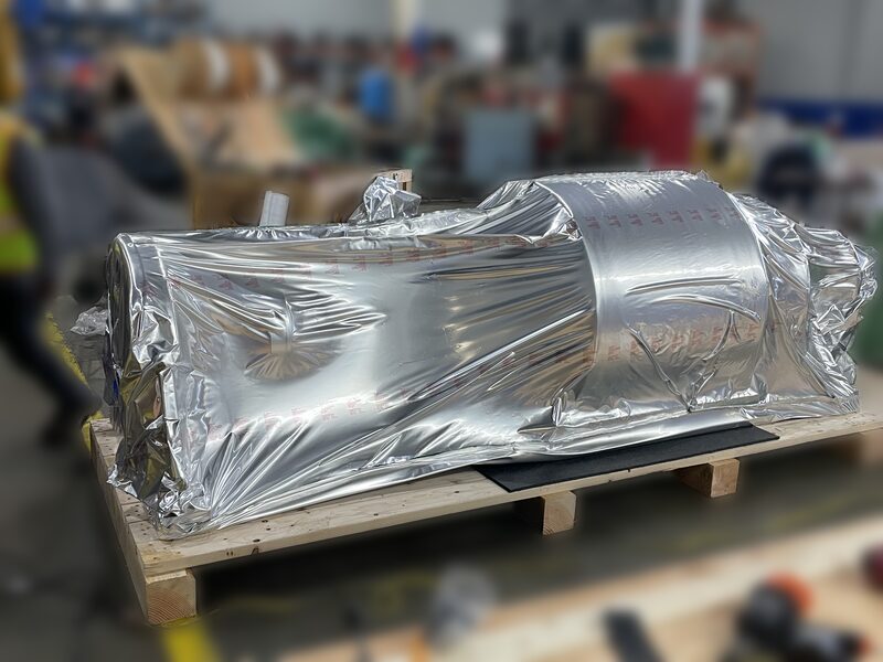 A machine sealed in a foil moisture barrier on a skid while its crate base is built alongside.