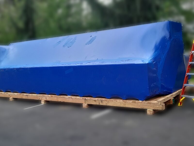 Blue industrial shrink wrap pulled tight over a long load on a timber skid outdoors.
